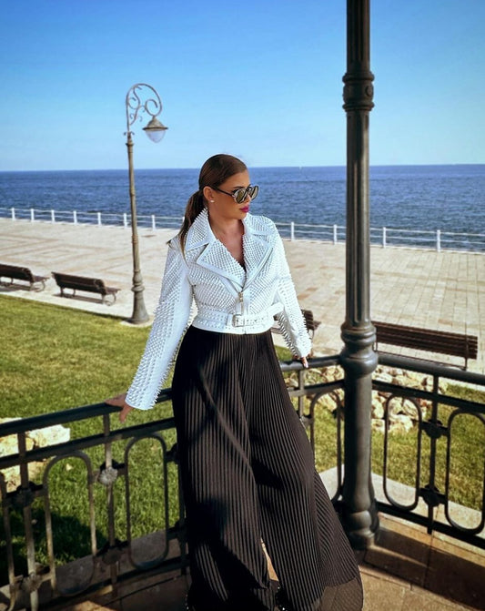 A model poses elegantly in the NICOLETTE white leather jacket by ROHA FURS, against a seaside backdrop.